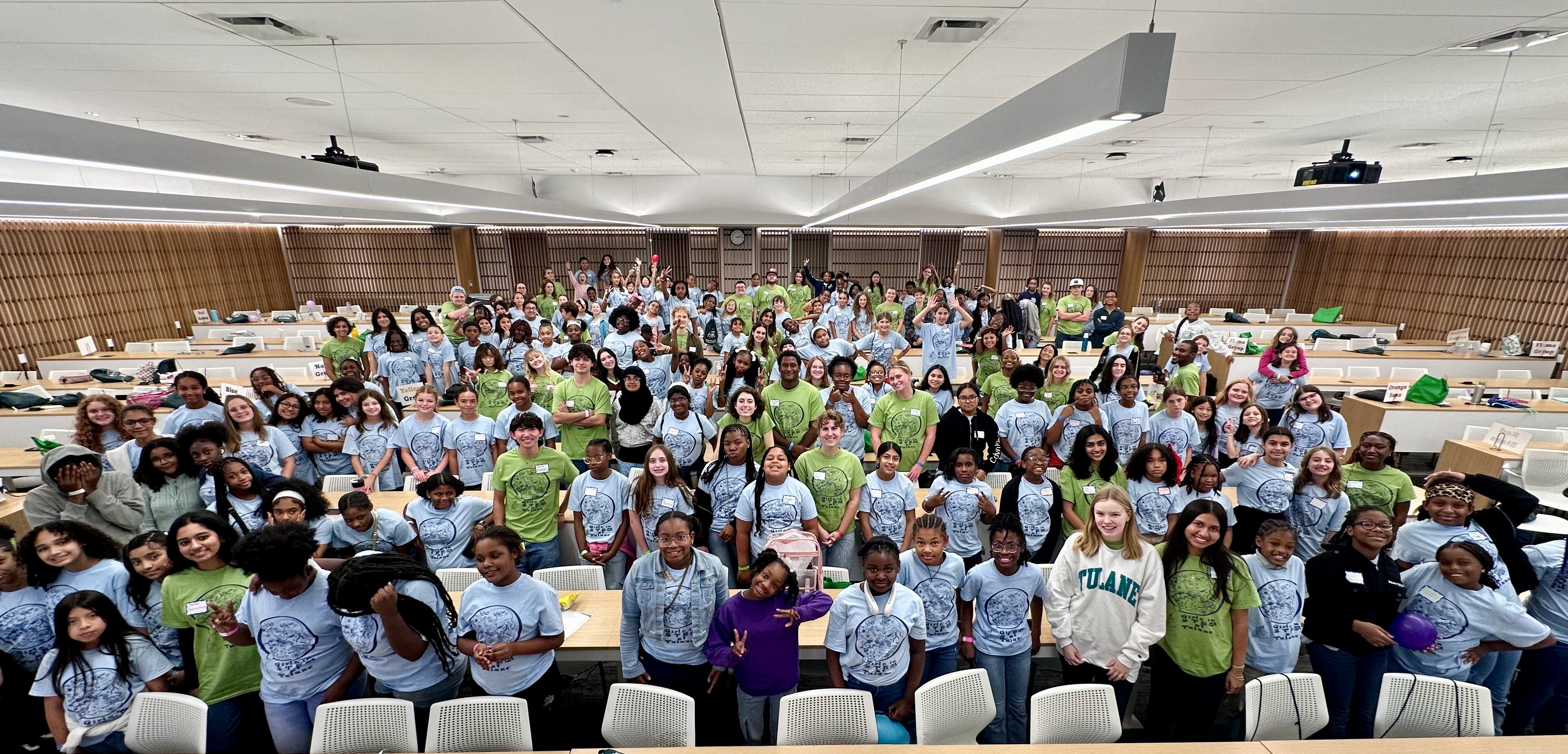 Large group of diverse students in blue and green shirts smiling in a lecture hall.