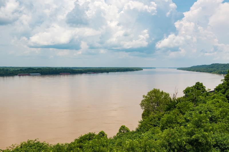 Wide, muddy river with barges, flanked by lush green trees under a cloudy sky.