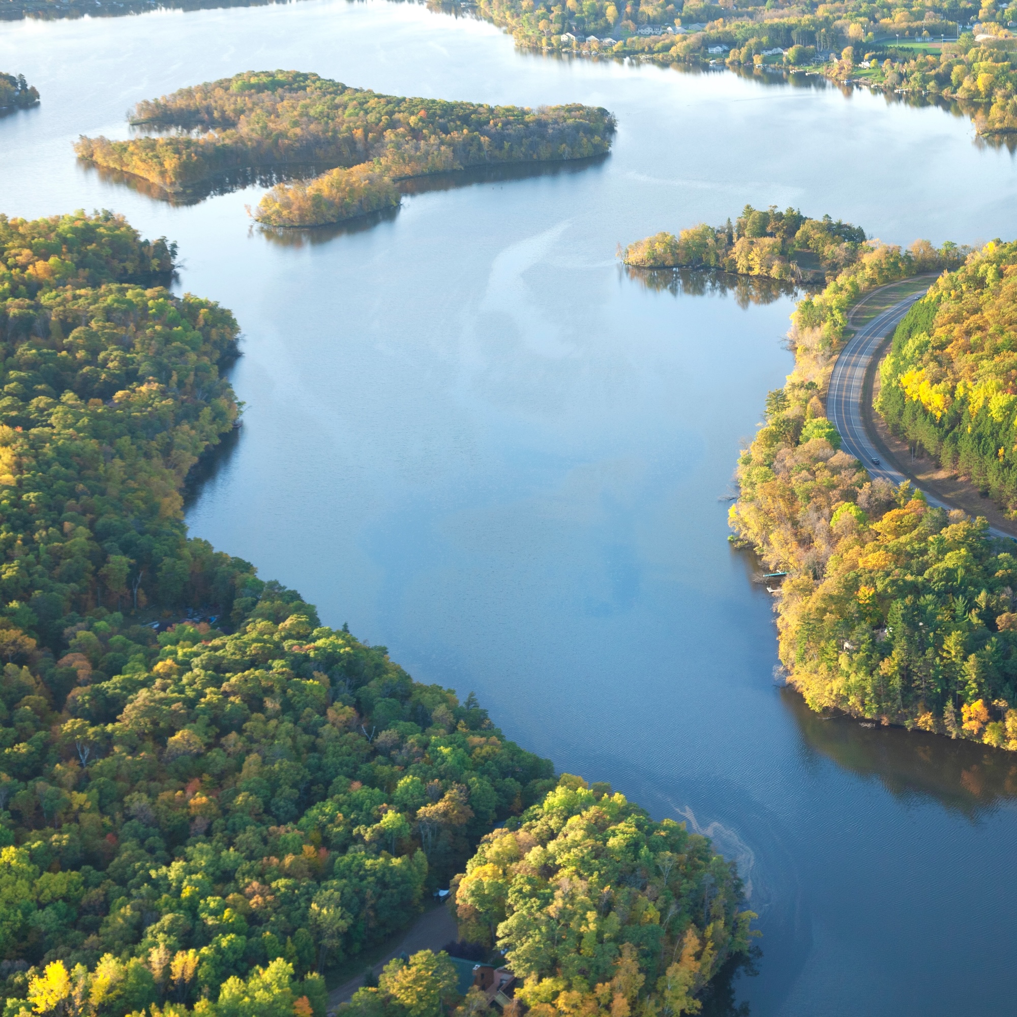 Aerial view of a lake with islands and autumn foliage, a road curves along the shore.