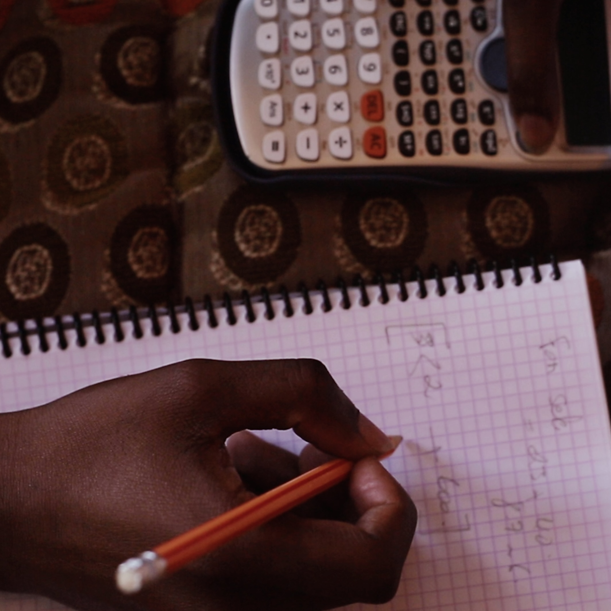 Close-up of a hand writing math problems in a notebook with a calculator nearby.