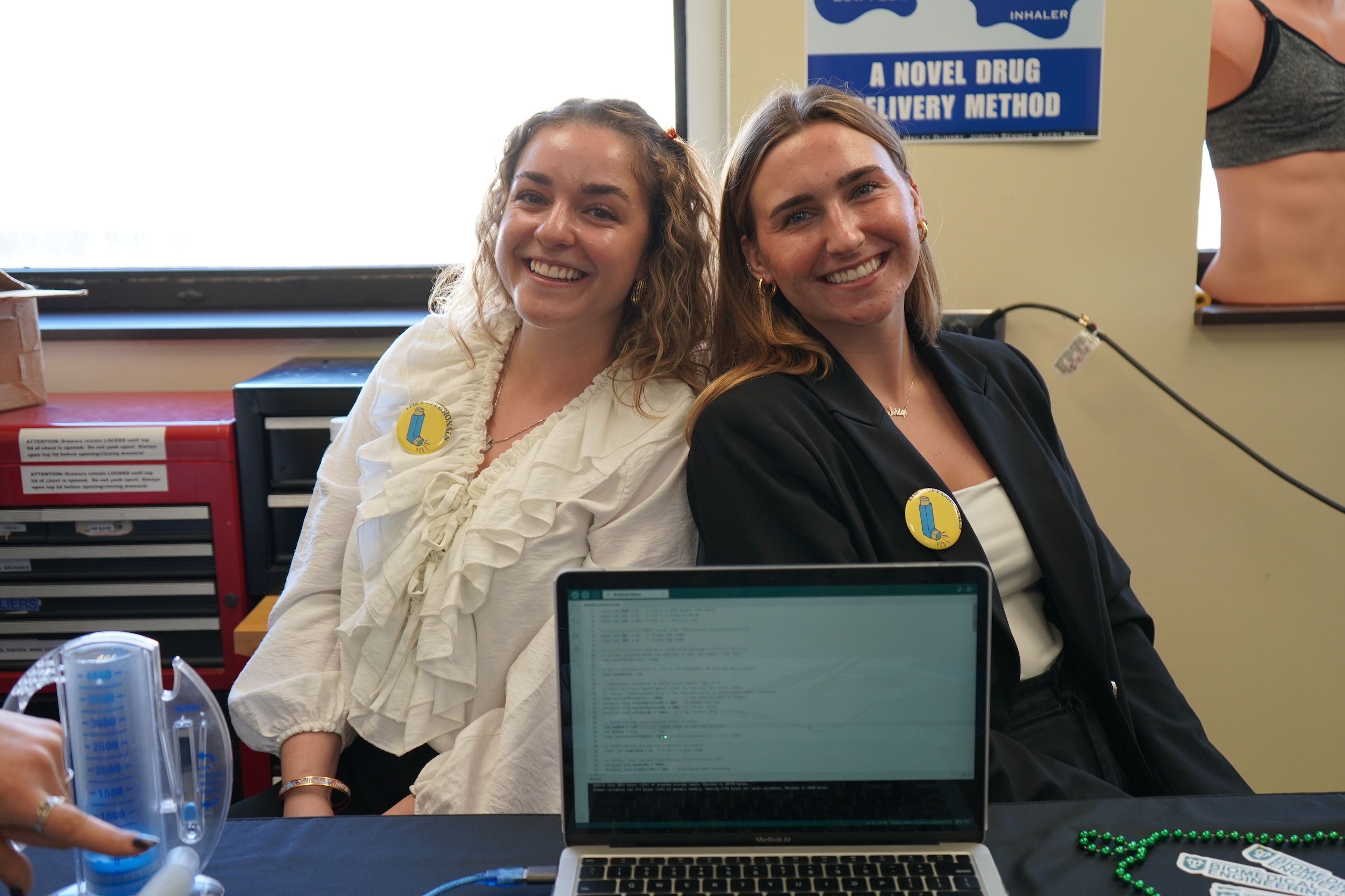 Two women smile at a table with a laptop, looking happy and approachable.