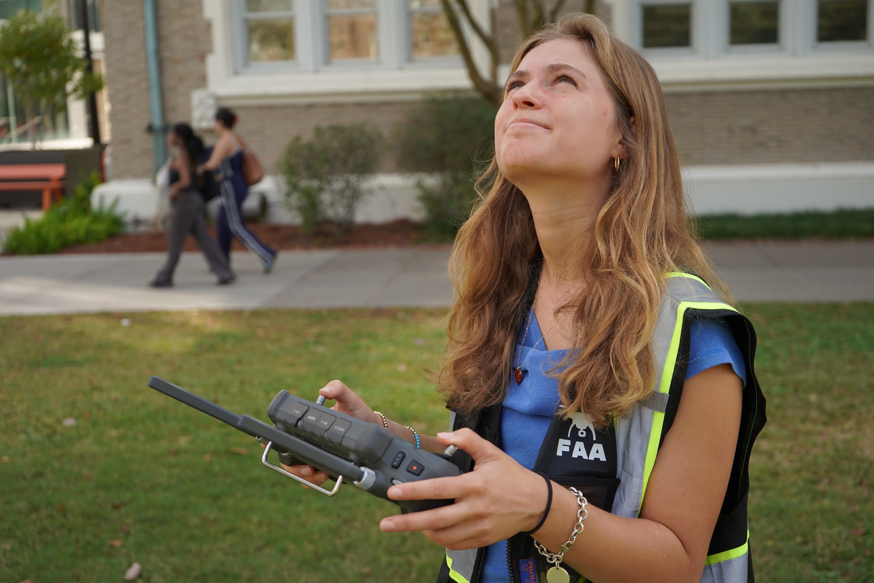 Young woman in a high-visibility vest holds a drone controller, looking up.