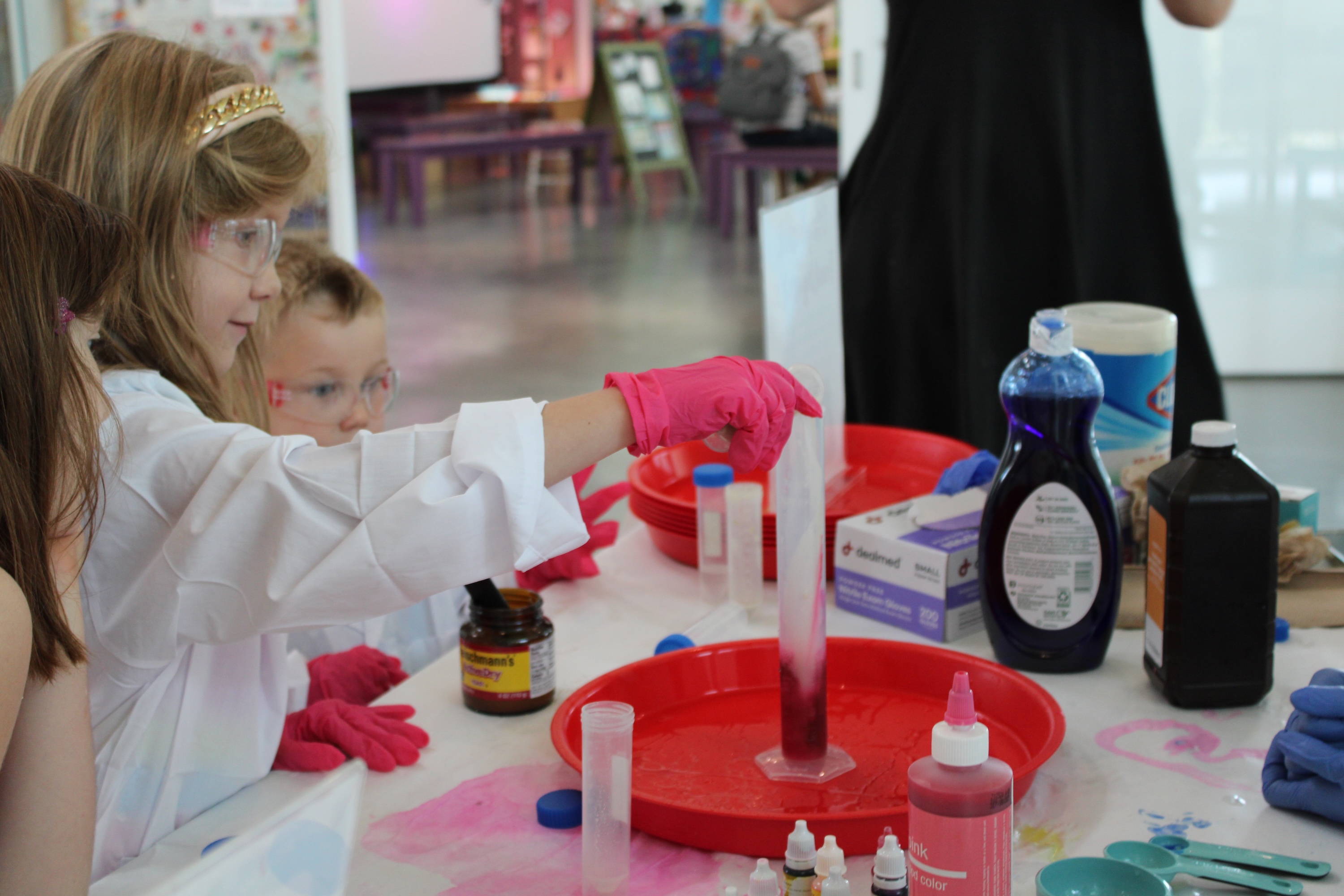 Child in lab coat and pink gloves conducts a science experiment with bubbling red liquid.