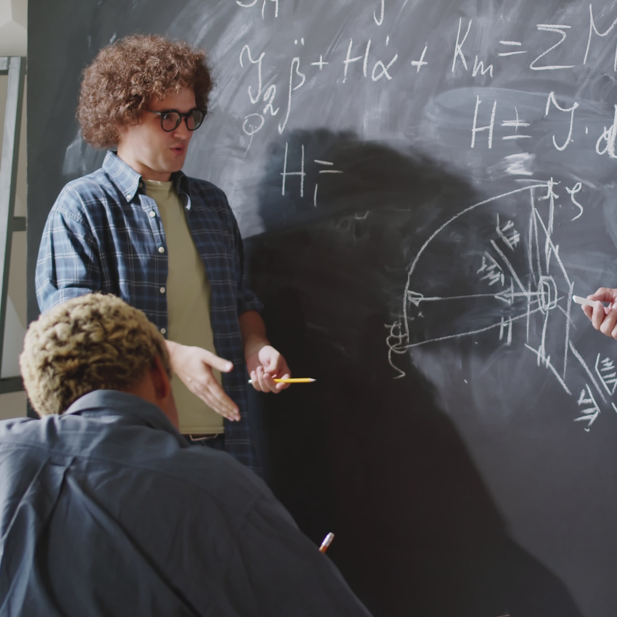 Man with curly hair explains math equations on a chalkboard to seated student.