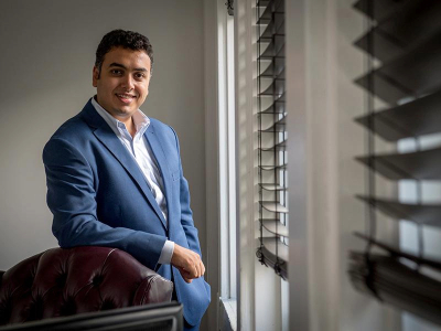 Michael Naguib in a blue suit leans on a leather chair next to a window.