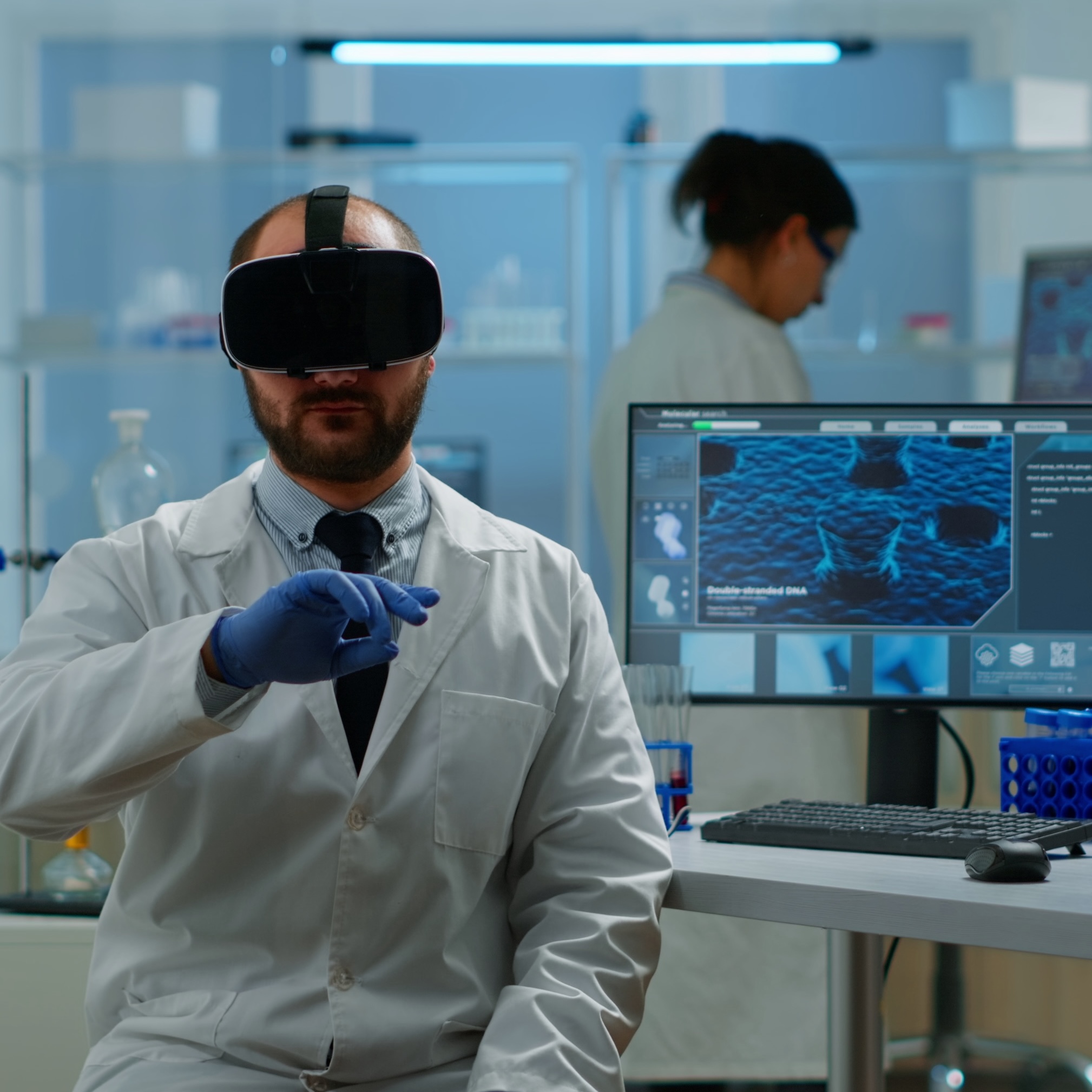 Scientist in VR headset gestures while looking at a cell culture on a monitor.