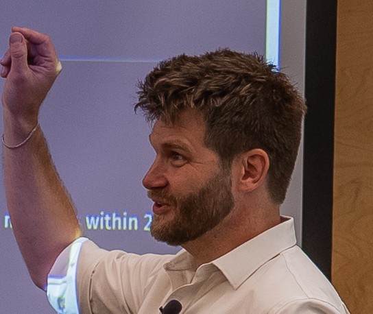 Man with beard and short hair, wearing a white shirt, gestures with his left hand raised.