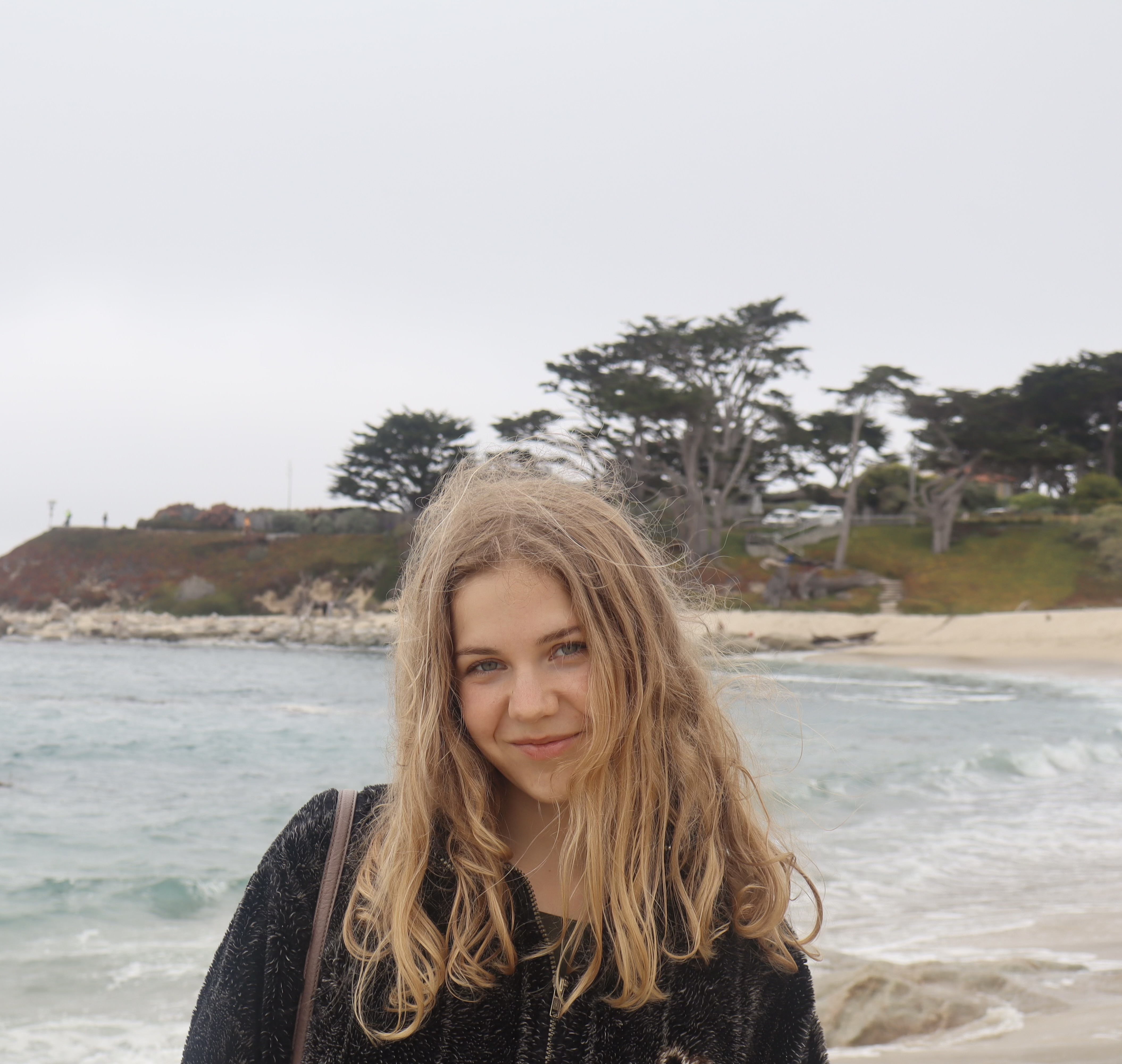 Young woman with long blond hair smiles at the camera on a sandy beach with trees in the background.