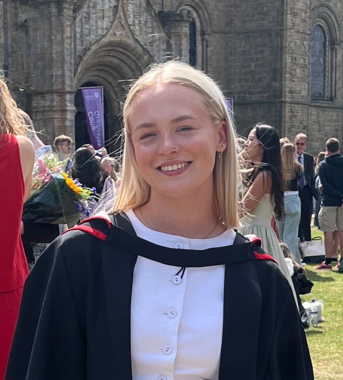 Smiling graduate in academic regalia in front of a stone building.