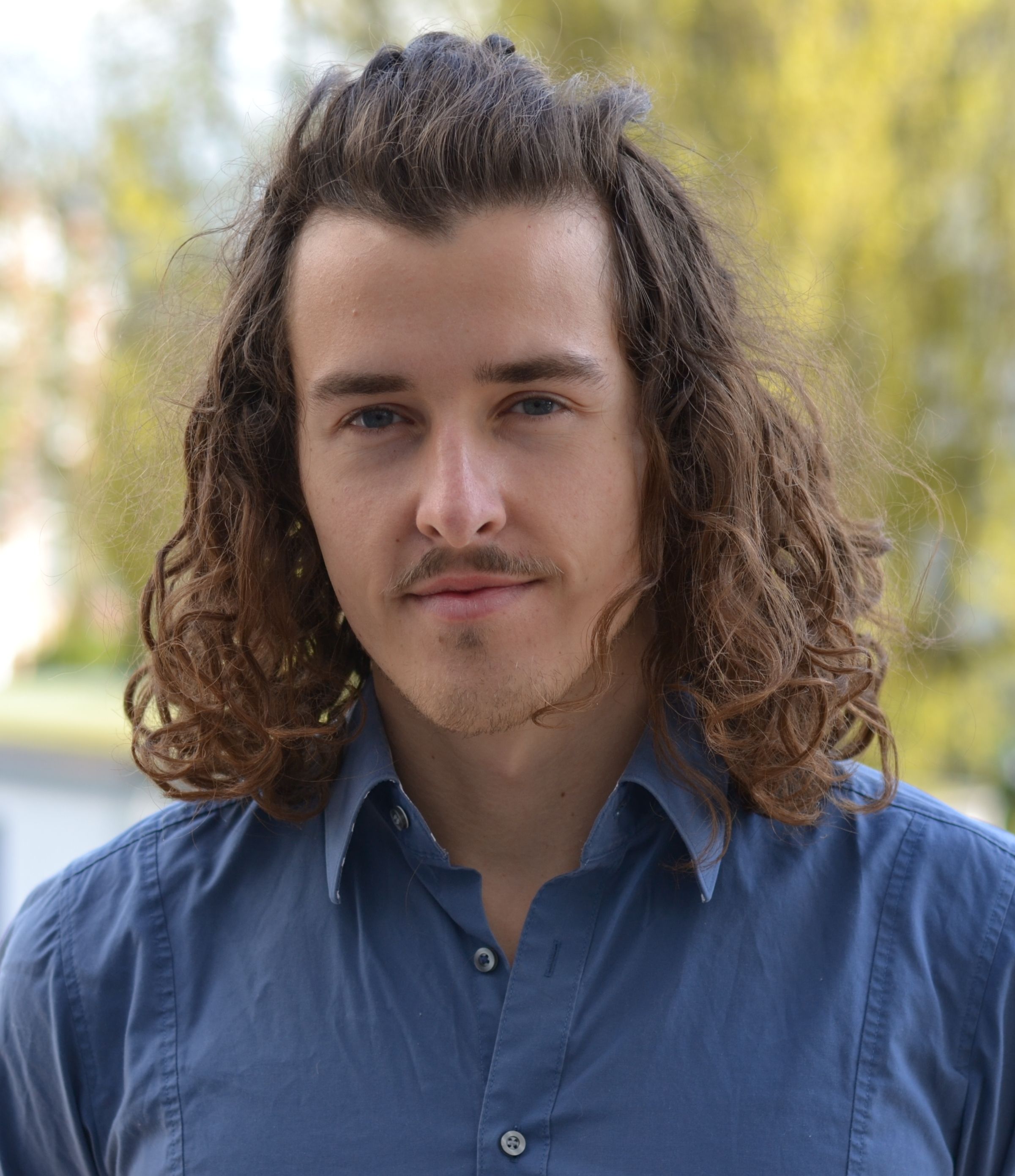 Young man with long curly brown hair and a mustache, wearing a blue collared shirt.