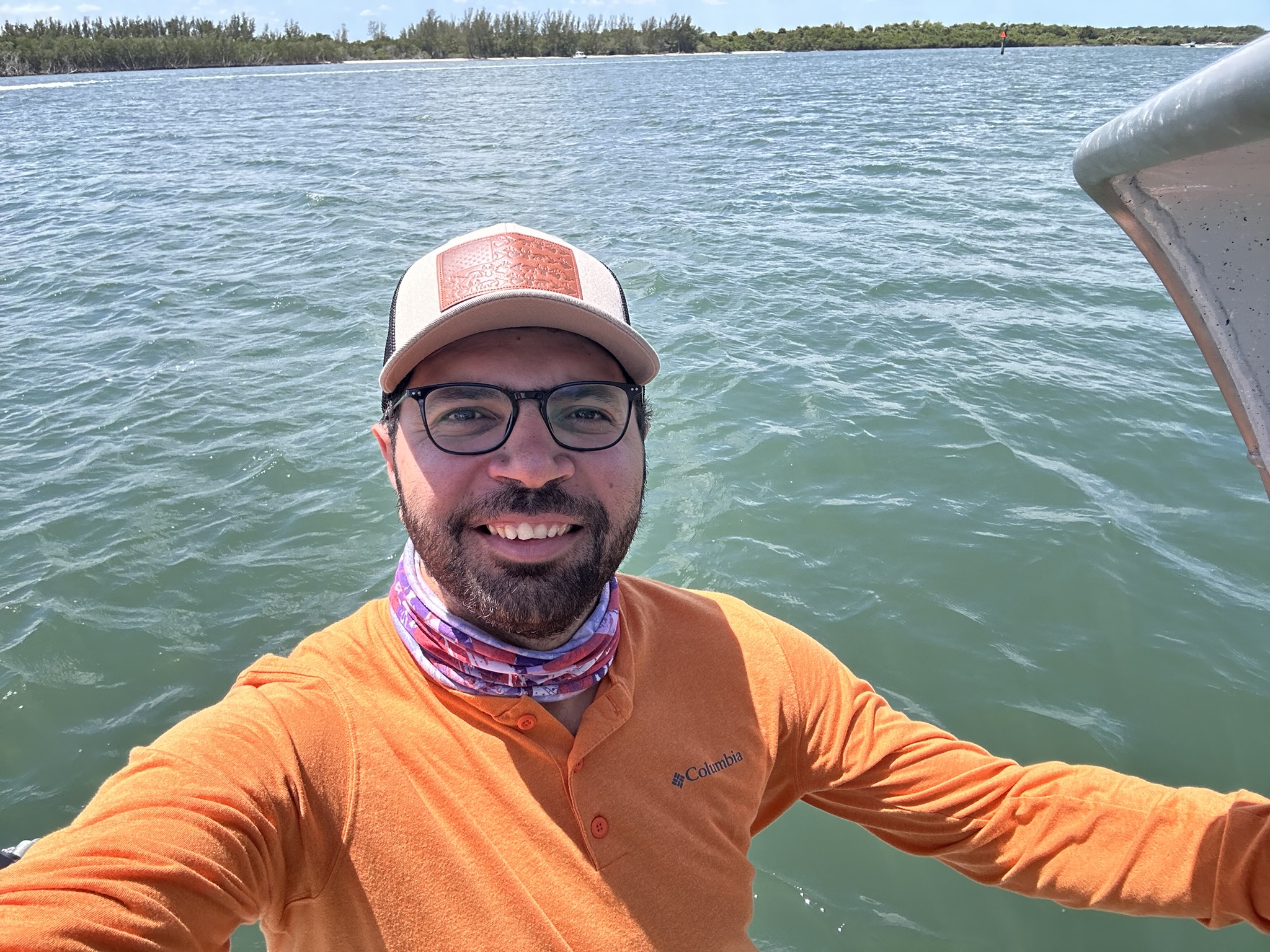 Man in orange shirt and hat smiles on a boat with water behind him.