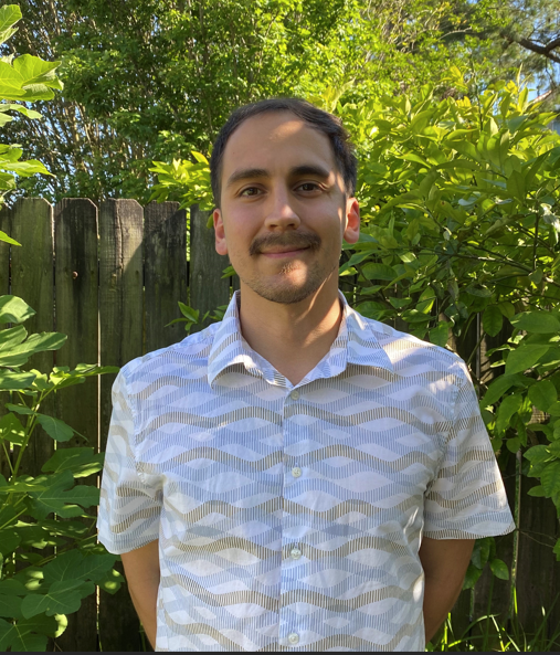 Man in a patterned shirt smiles in a garden, with a wooden fence behind him.
