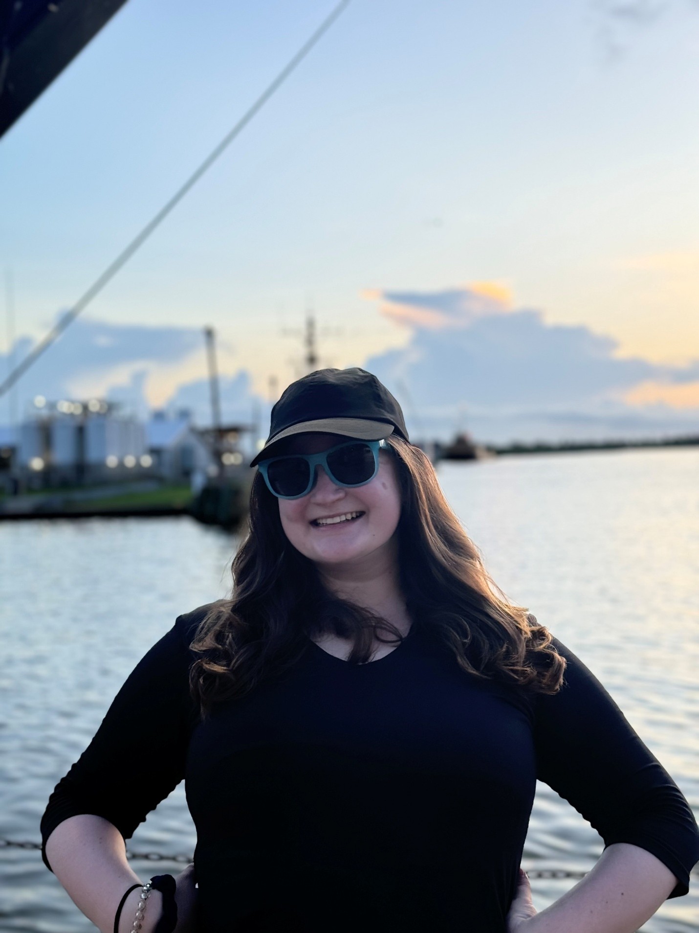 Young woman in baseball cap and sunglasses smiling at sunset by the water.