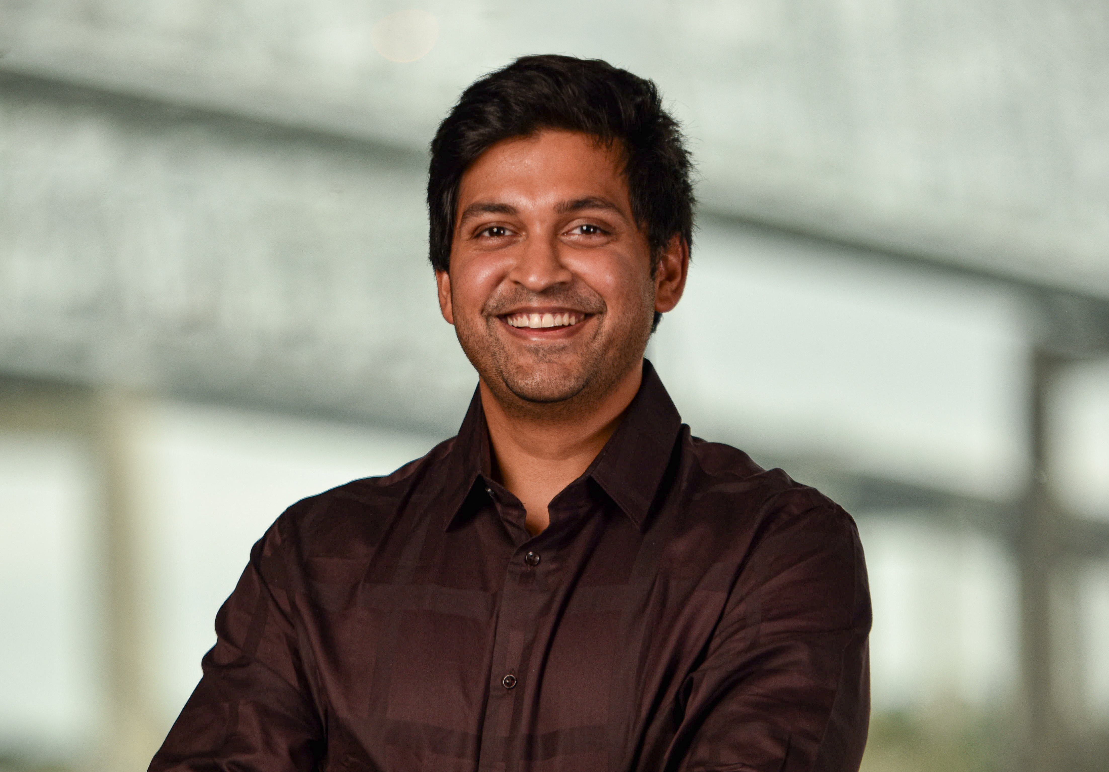 A man with dark hair, smiling, wearing a dark collared shirt.