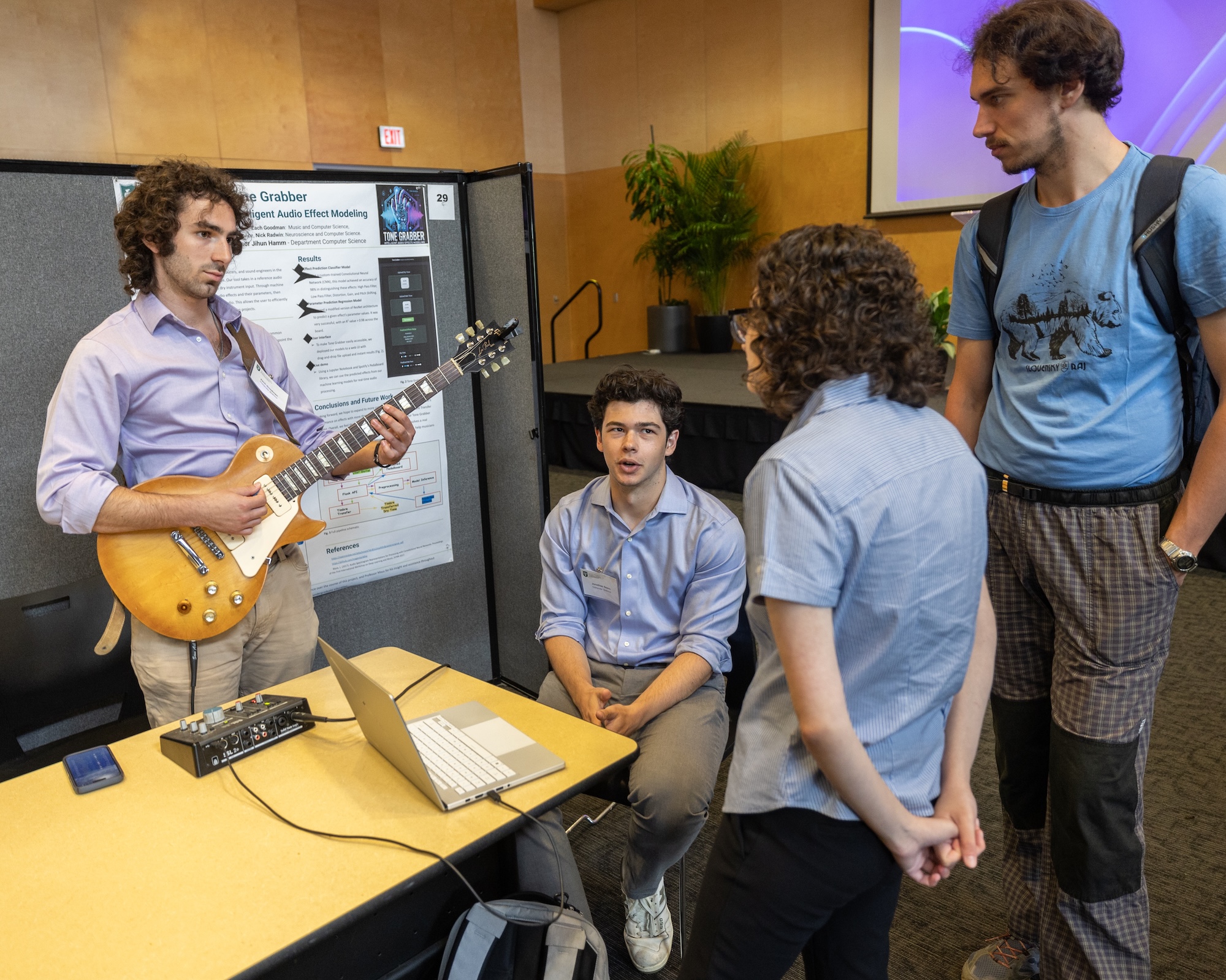 Student plays guitar near a poster board while three others watch.