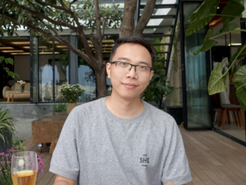 Young man in a grey t-shirt and glasses smiles at the camera, outdoors.