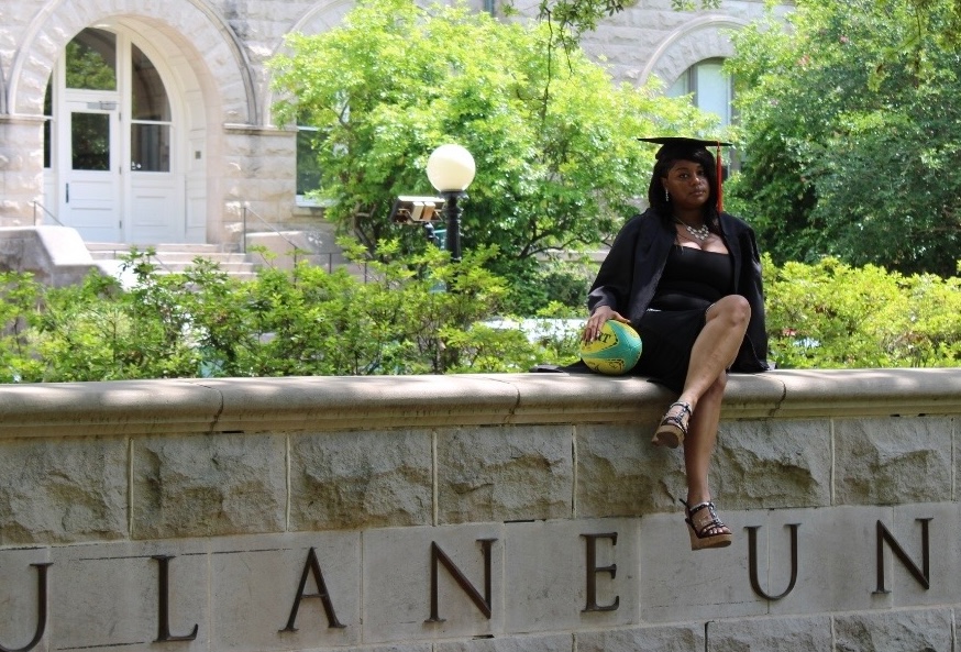 Woman in graduation cap and gown sits on stone wall with "TULANE UN..." carved into it.