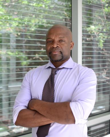 A bald Black man in a light purple shirt and patterned tie with arms crossed stands in front of a window with blinds and greenery.