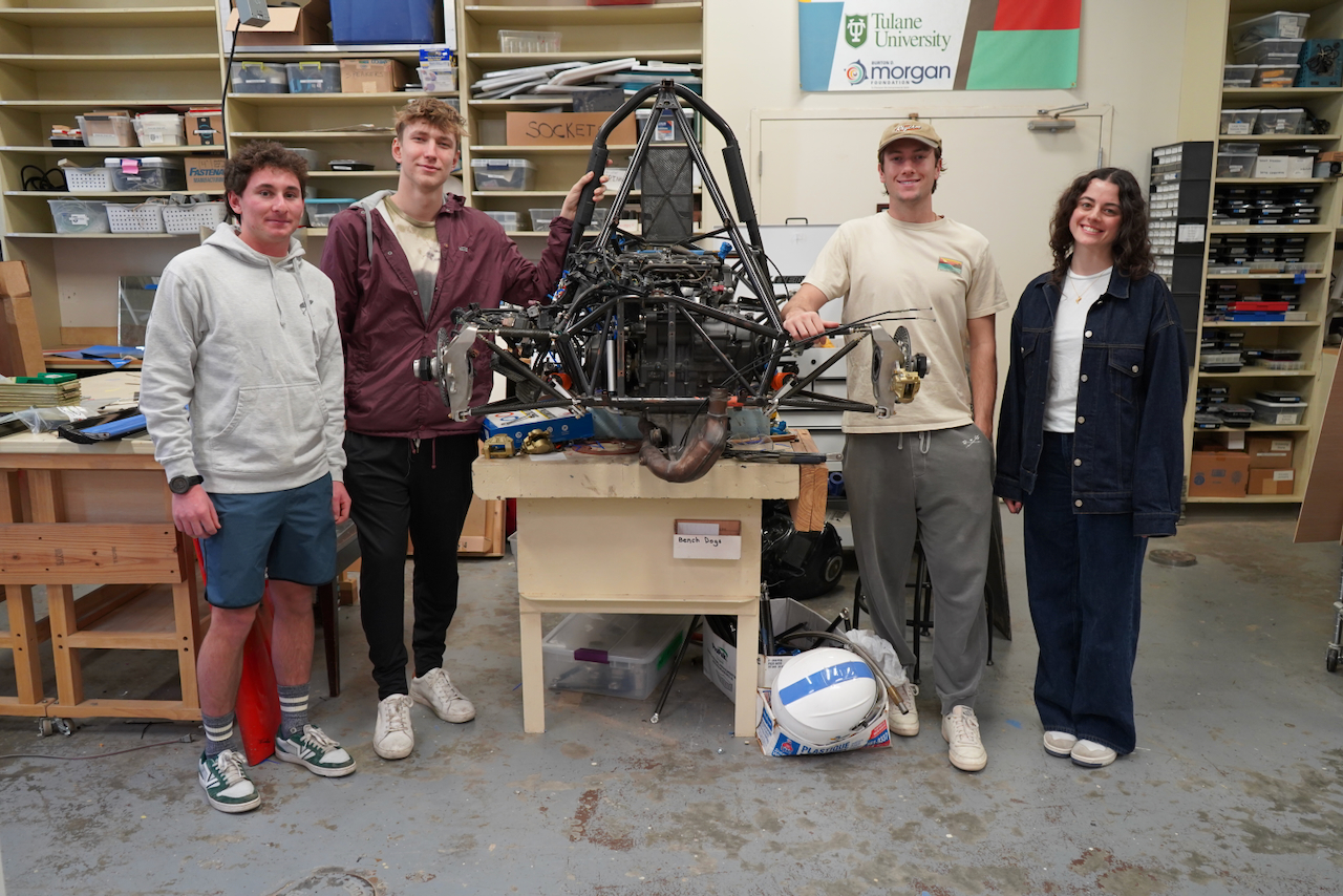 Four smiling students stand around a large, complex machine in a workshop.