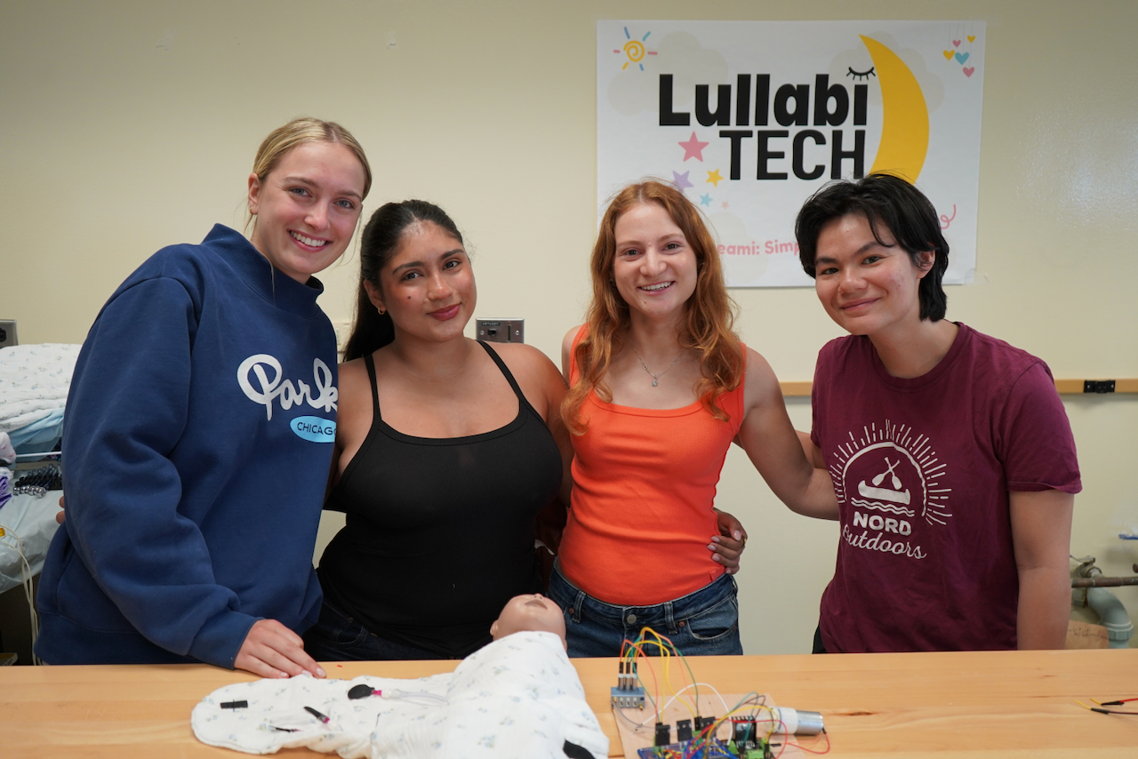 Four smiling young women pose for a photo behind a table with a baby doll and tech equipment.
