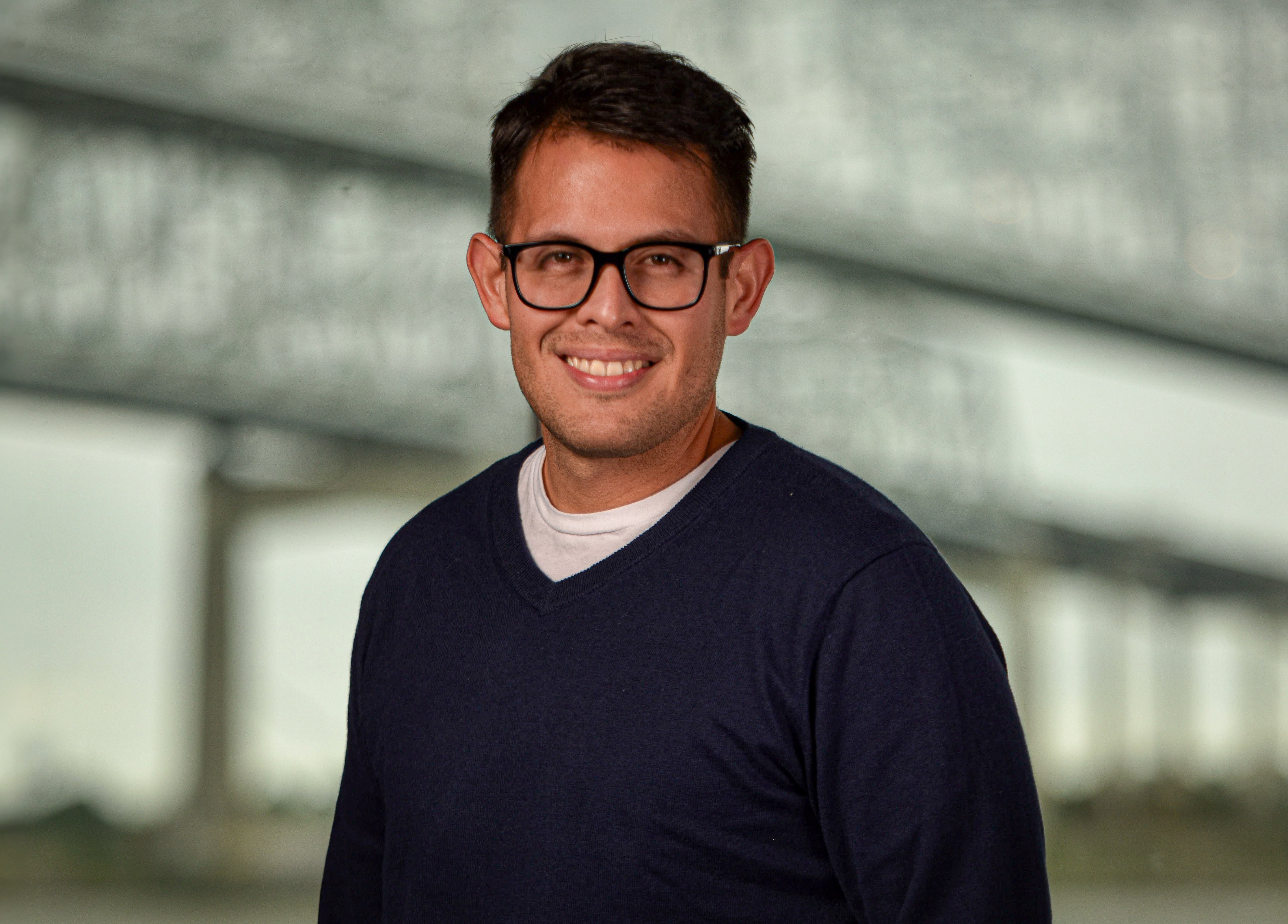A man wearing glasses and a navy blue v-neck sweater over a white t-shirt smiles at the camera.