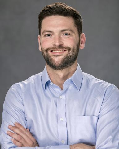 A man with a beard and blue shirt smiles with arms crossed.