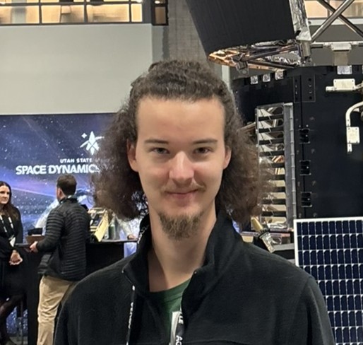 Young man with curly brown hair and a goatee, wearing a black jacket, smiles at the camera.