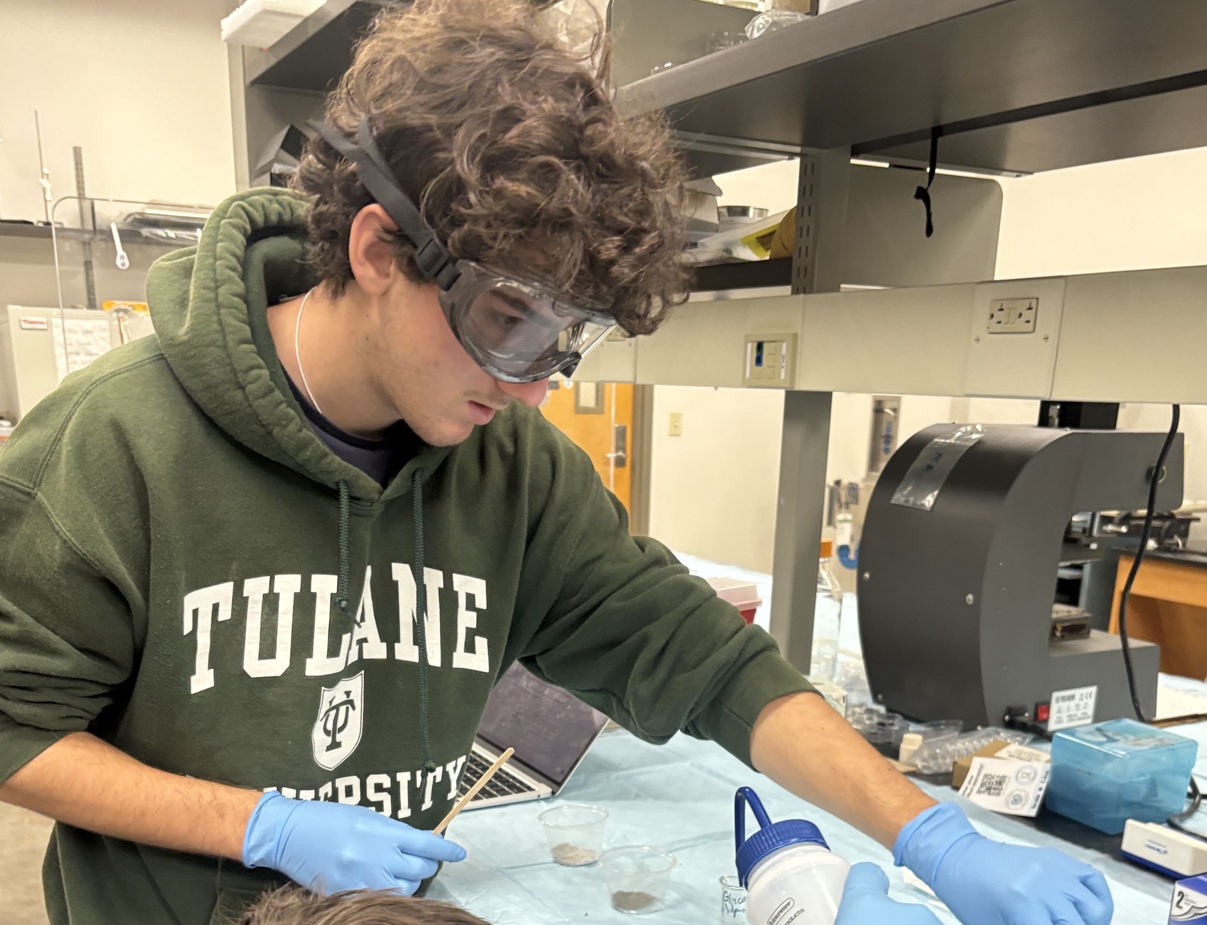 Student in a Tulane University hoodie and safety goggles works at a lab bench.