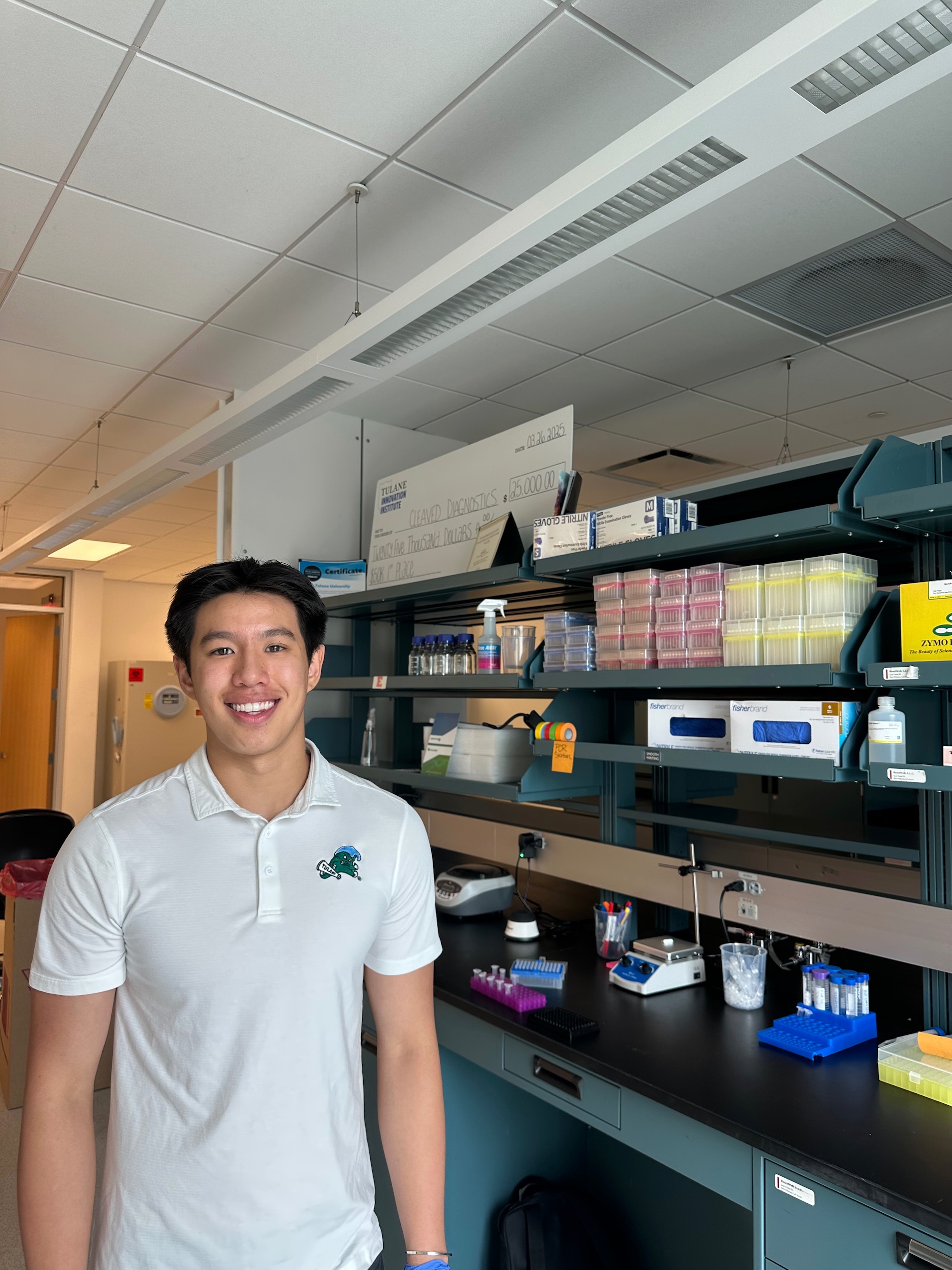 A young man in a white polo shirt smiles in a laboratory with shelves of supplies.
