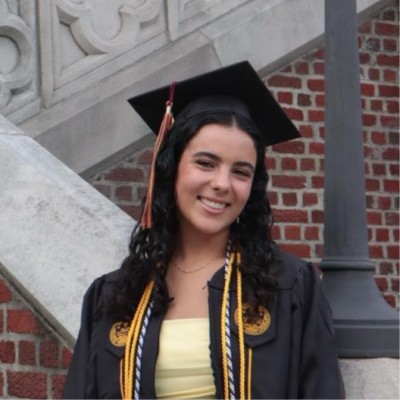 Young woman in graduation cap and gown, smiling proudly outdoors.