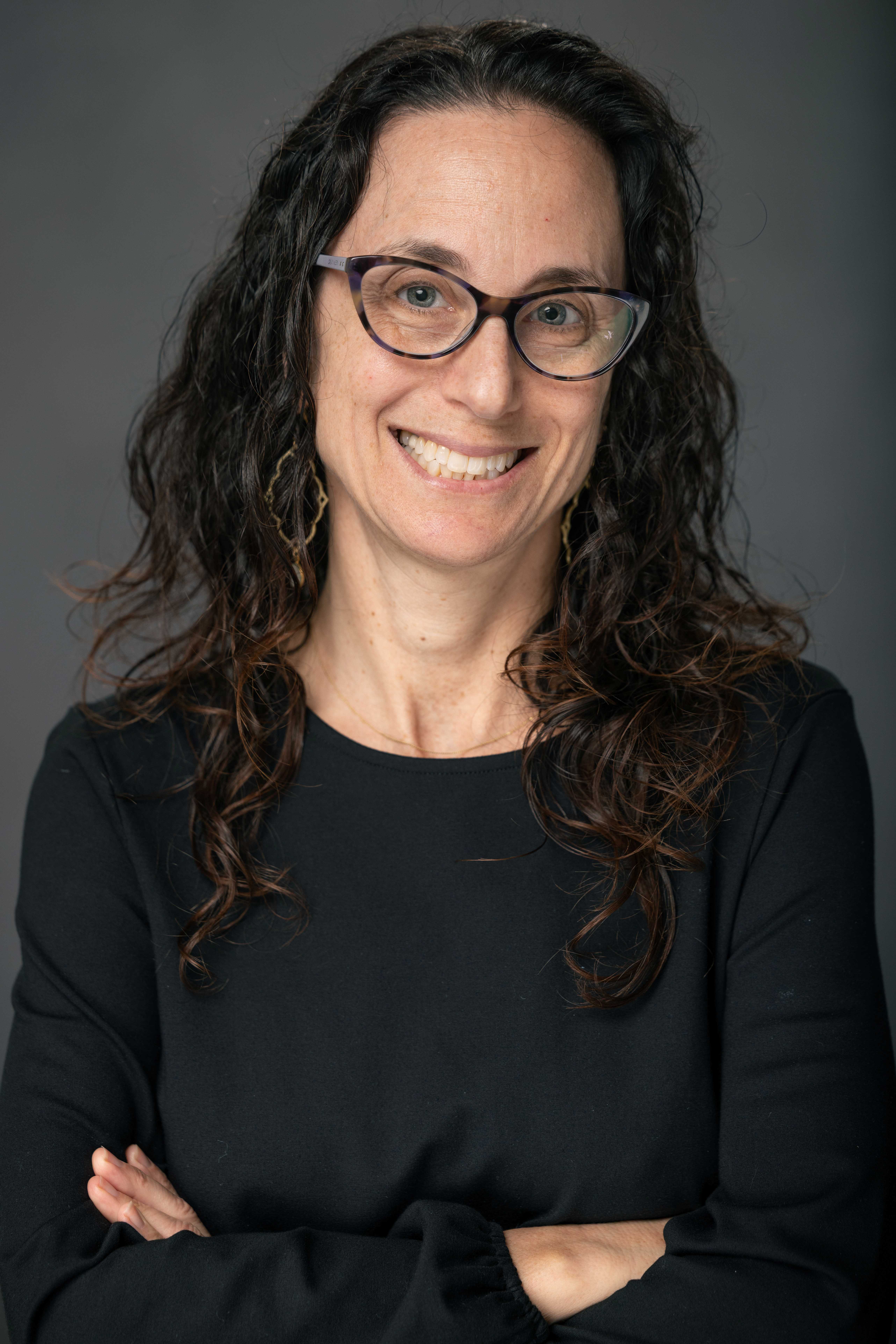 Smiling woman with dark curly hair and glasses, arms crossed.
