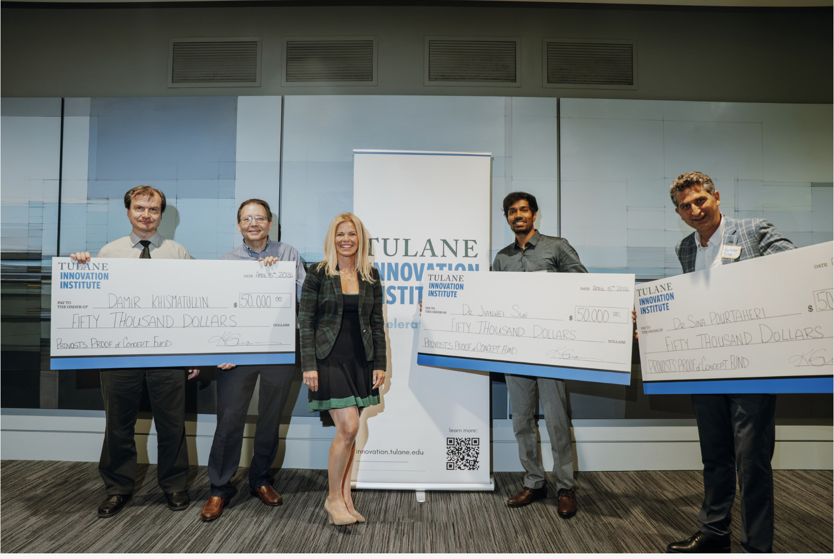 Five people smile while holding large checks in front of a banner.