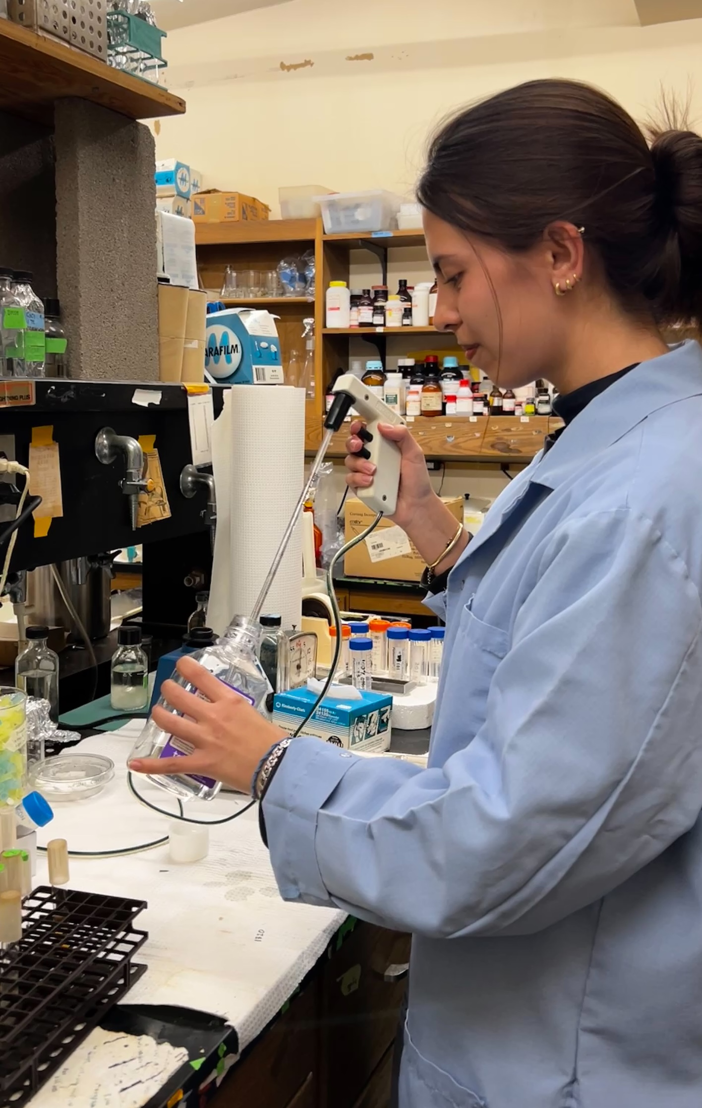 A woman in a lab coat uses a pipette to transfer liquid into a flask.