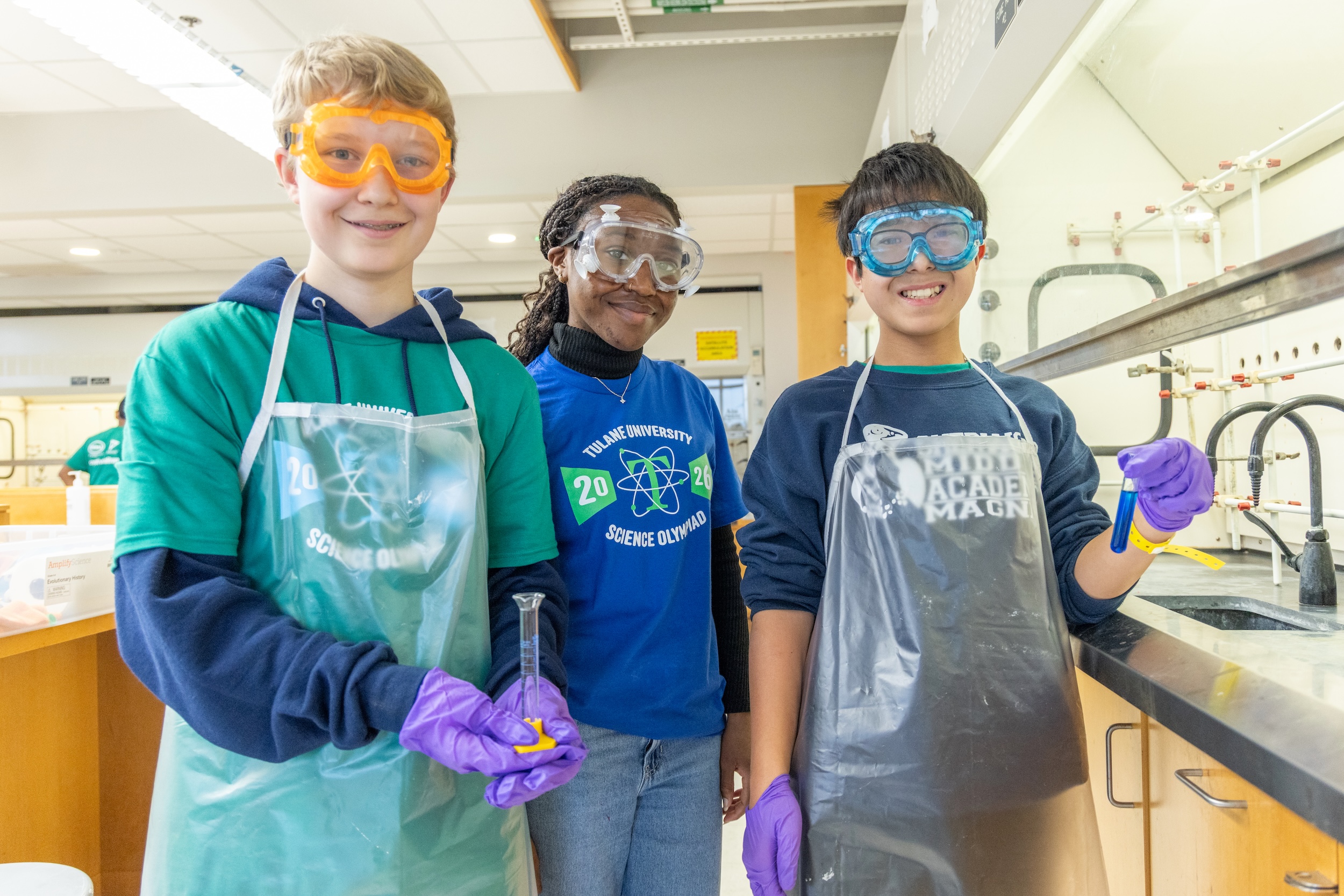 Three smiling children in a lab, wearing goggles and aprons, holding test tubes.