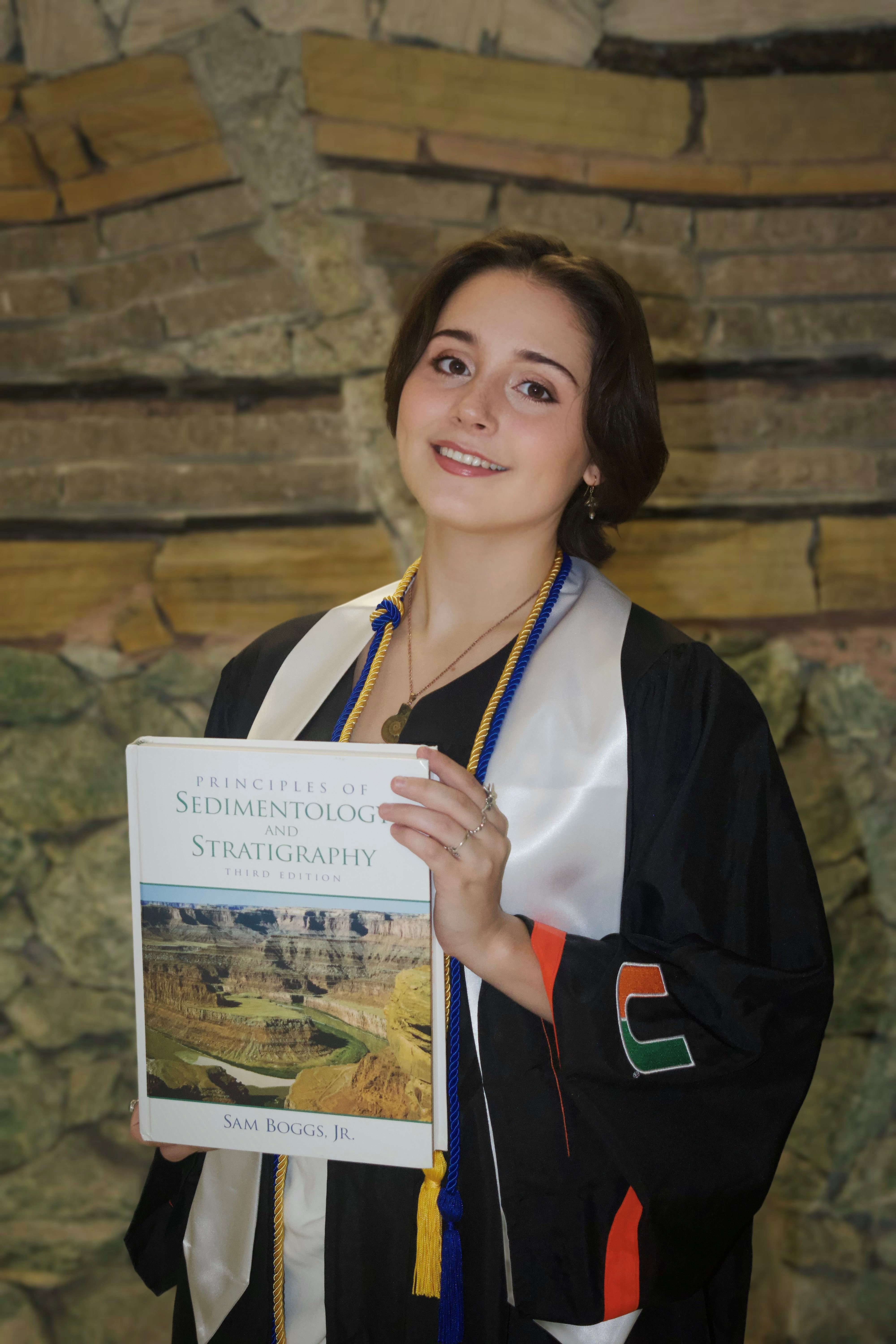 Graduating student in cap and gown holds up a book titled "Stratigraphy".