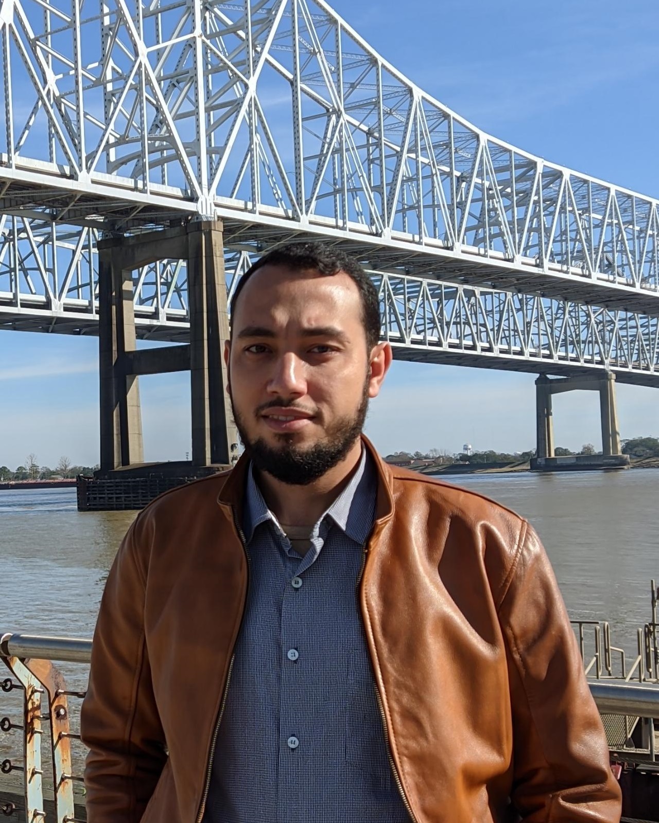 Man in brown jacket in front of a large bridge.