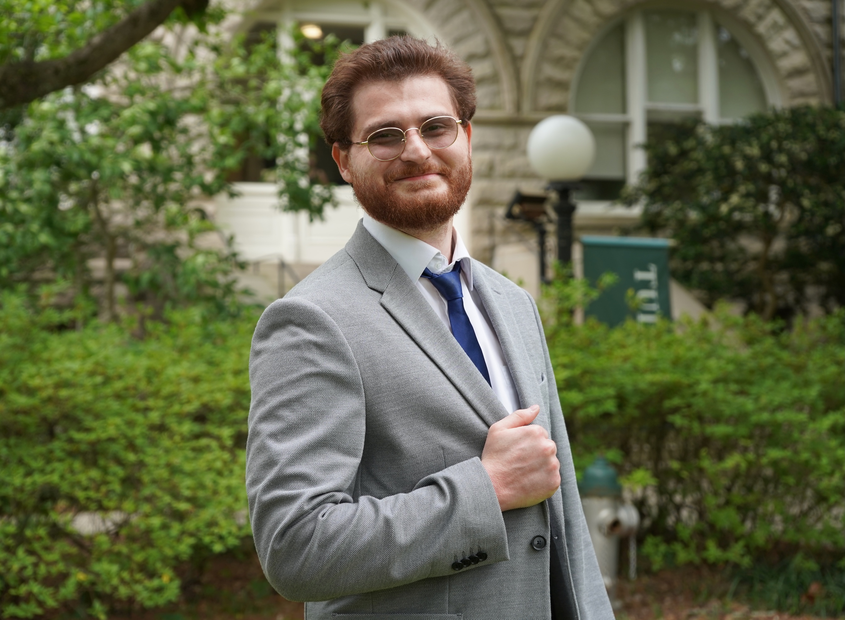 Man in a suit, smiling, with a building and greenery behind him.