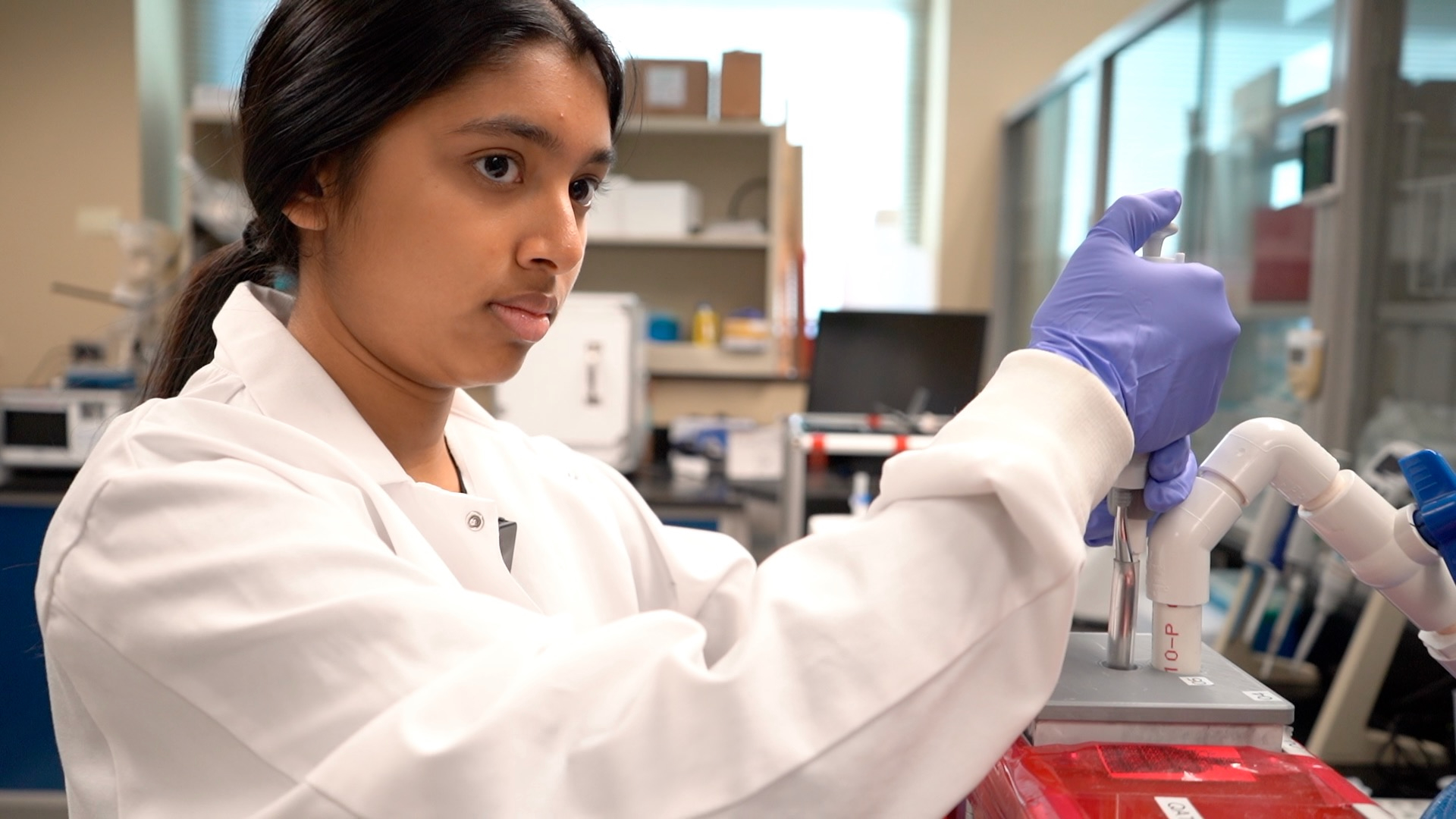 A young woman in a lab coat and gloves uses a pipette.