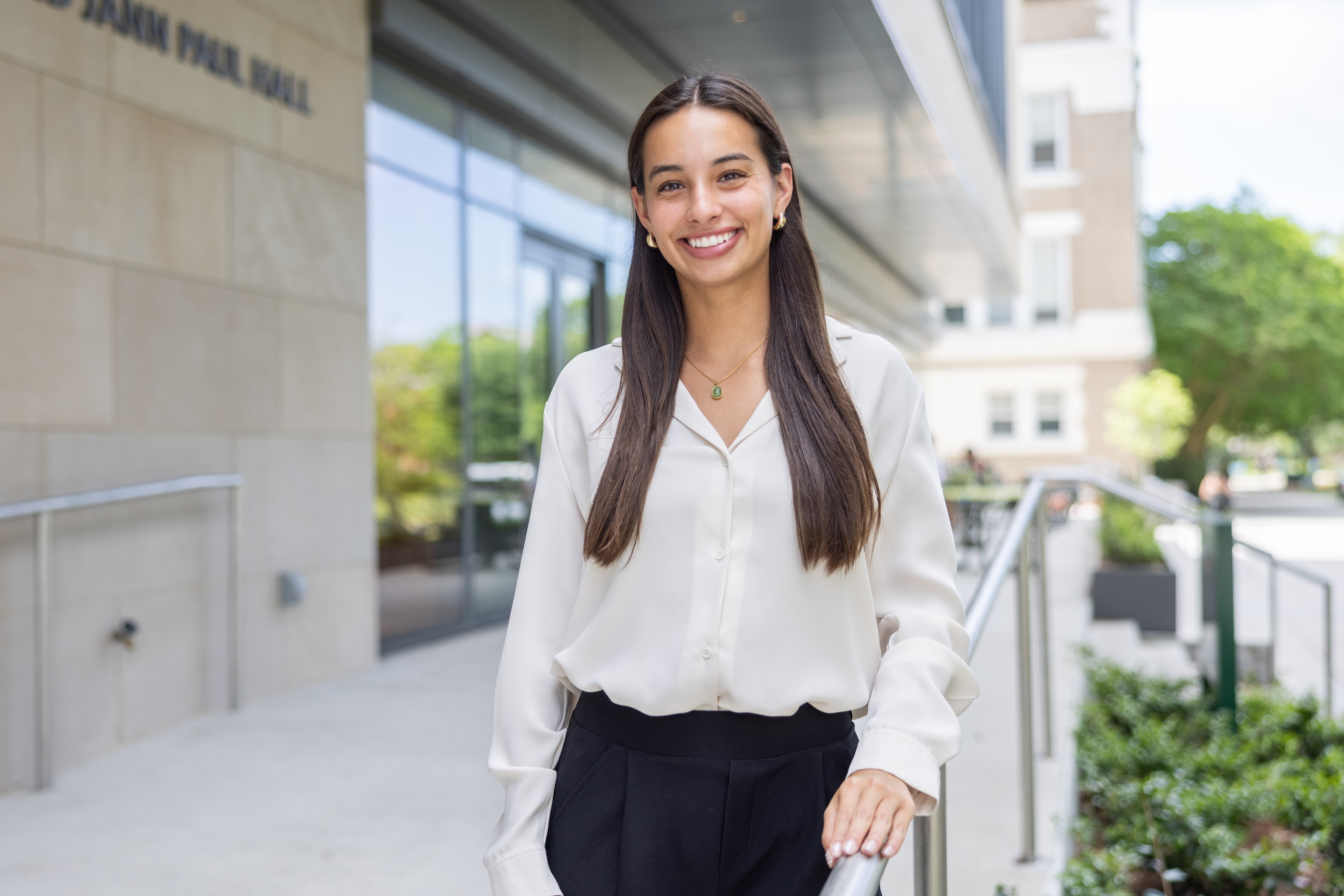 Young woman in white blouse and black pants smiles outside a building.