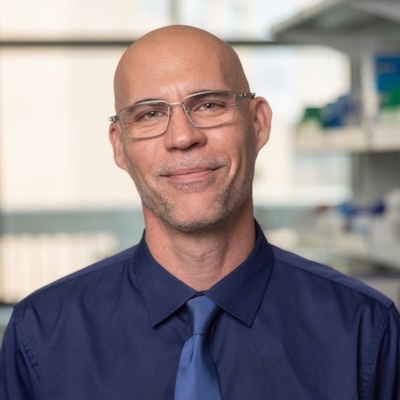 Man in glasses with dark blue shirt and light blue tie smiling at camera