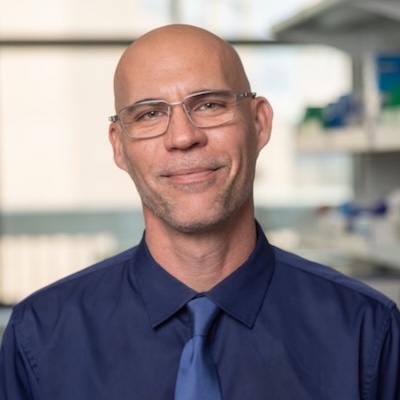 Man in glasses with dark blue shirt and light blue tie smiling at camera