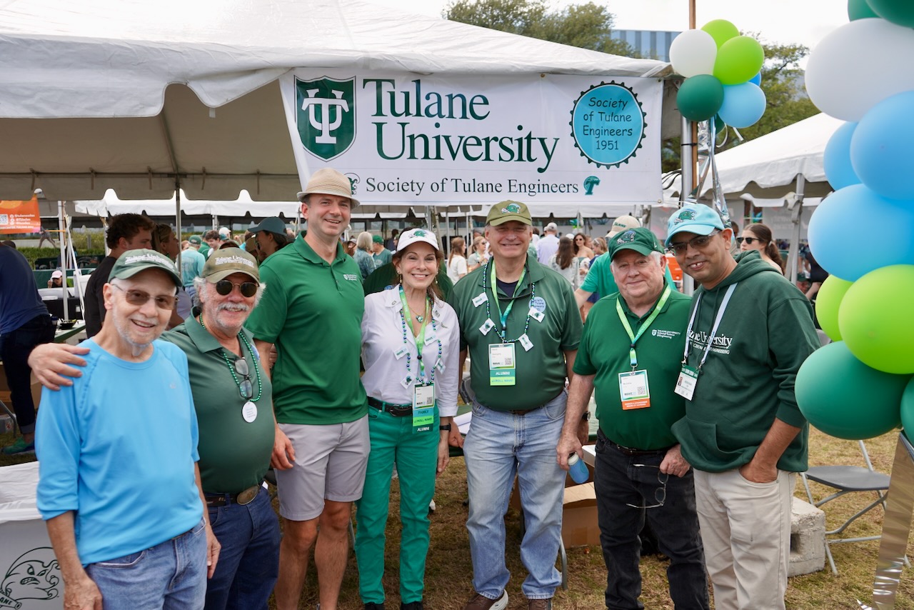 Group of people in blue and green smiling in front of tailgating tent
