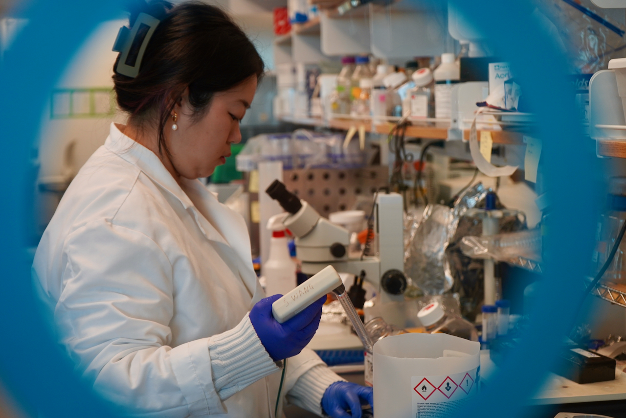 Scientist in lab coat and blue gloves examines a device in a laboratory, framed by a blue circle.