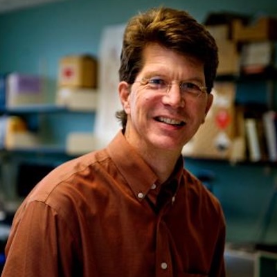 A man wearing glasses and a brown button-down shirt smiles.or