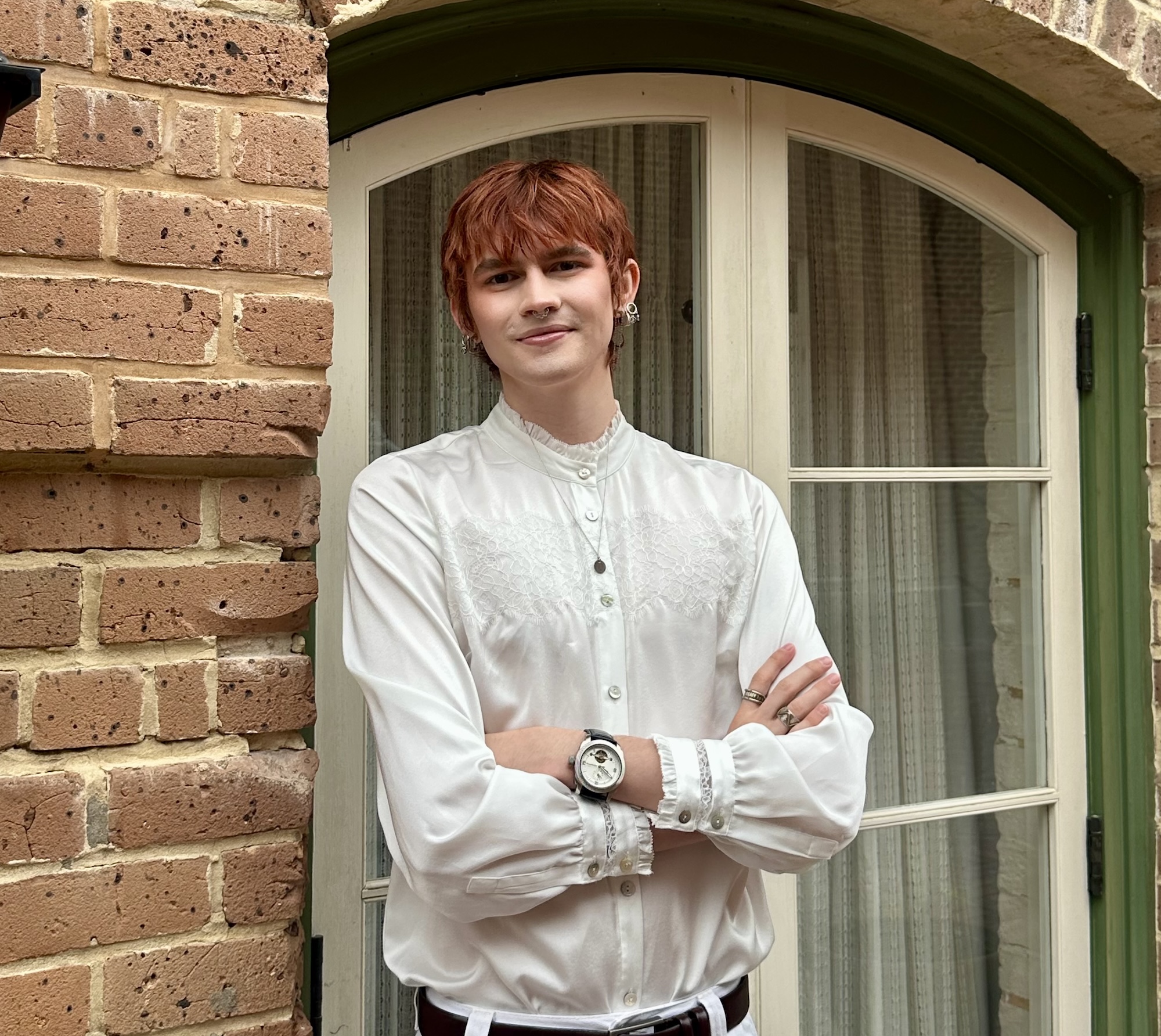 Person with red hair, wearing a white ruffled shirt, standing with arms crossed.