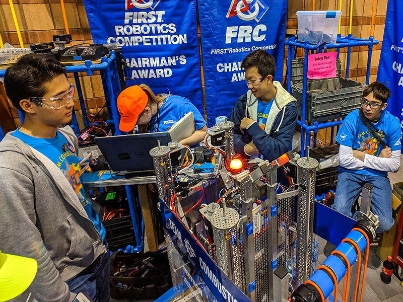 Young people in safety glasses work on a complex robot at a robotics competition.