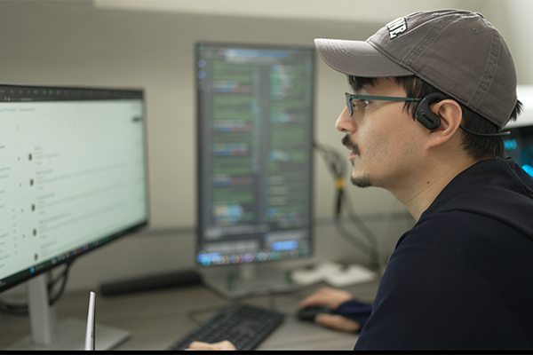 Man in a baseball cap and glasses works at two computer monitors.