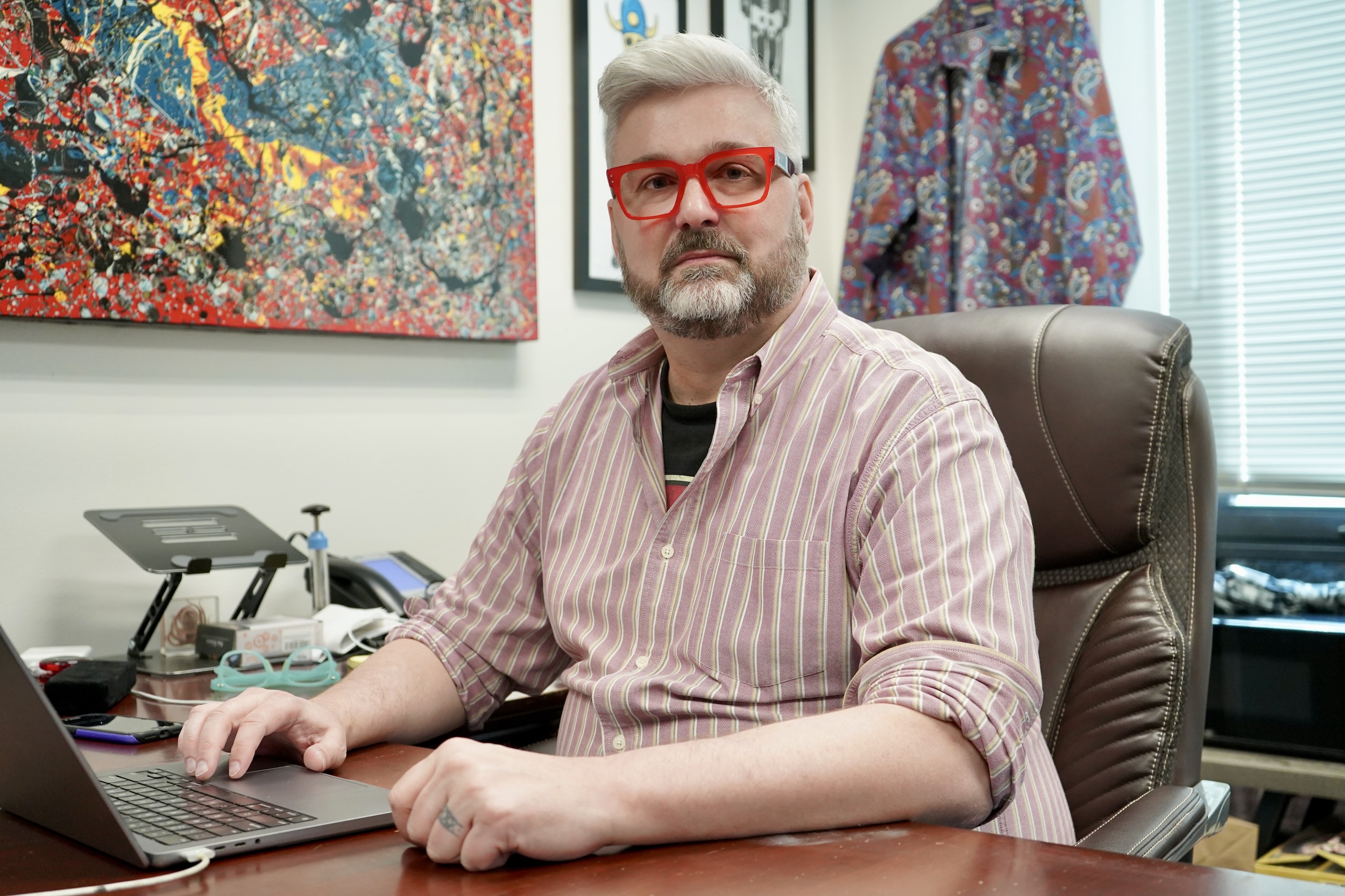 A man with gray hair, a beard, and red glasses sits at a desk in an office, looking at the camera.