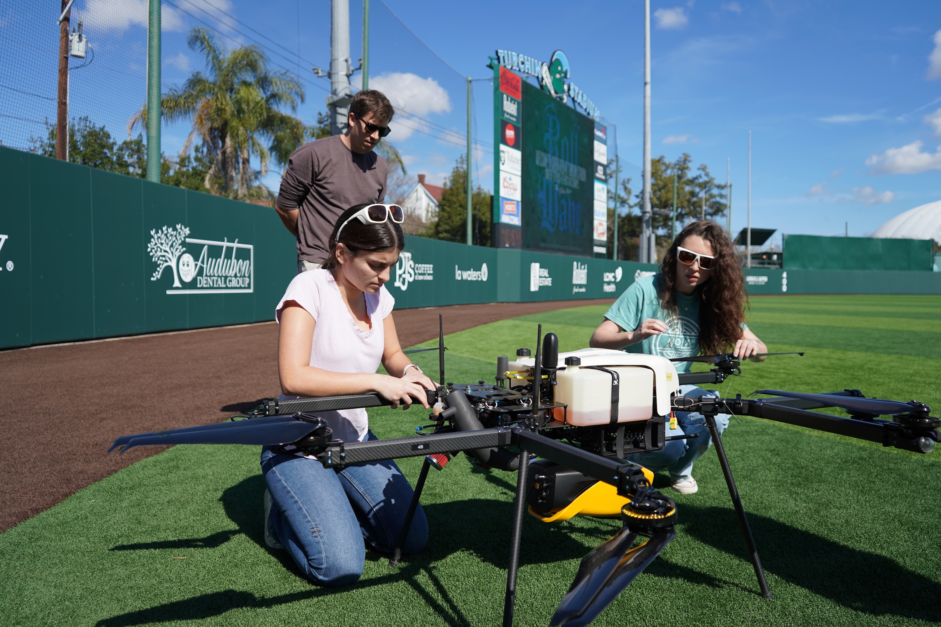 Three people examine a large drone on a sunny baseball field.