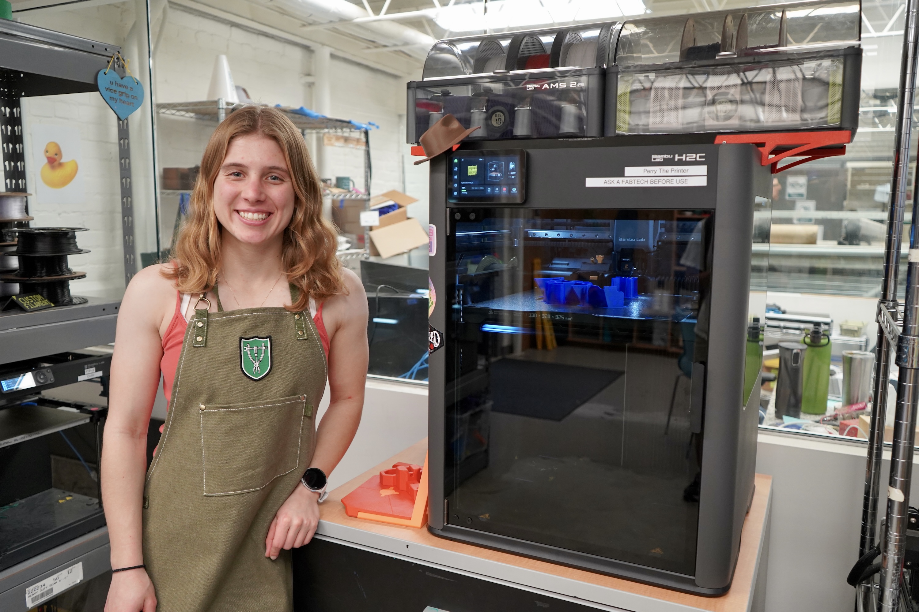 Young woman in apron smiles next to a large 3D printer.